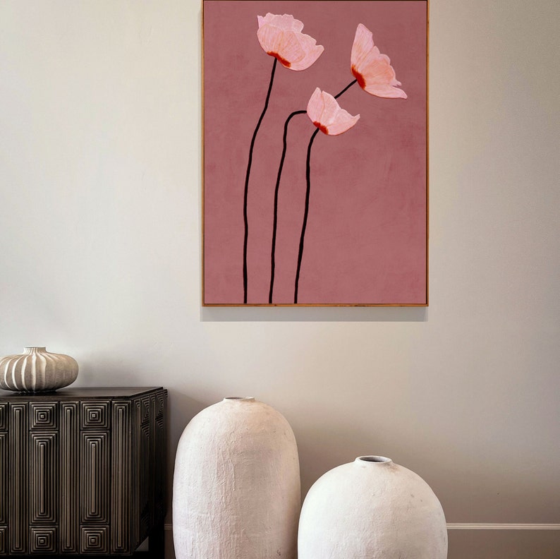 May include: A framed floral art print featuring three pink flowers with black stems against a dusty rose background. The artwork is in a gold frame and is displayed on a white wall. The style is minimalist and modern.