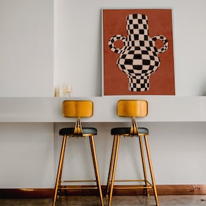 May include: Two gold bar stools with black leather seats sit in front of a white counter. A framed print of a black and white checkered vase on a red background hangs on the wall behind the counter.