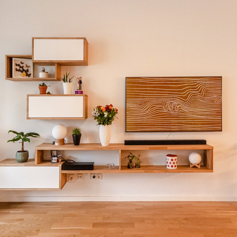 May include: A white and brown wooden shelf unit with a TV mounted on the wall above it. The shelf unit has a variety of items on it, including plants, a vase of flowers, and a decorative object. The TV is a flat screen and is turned off.