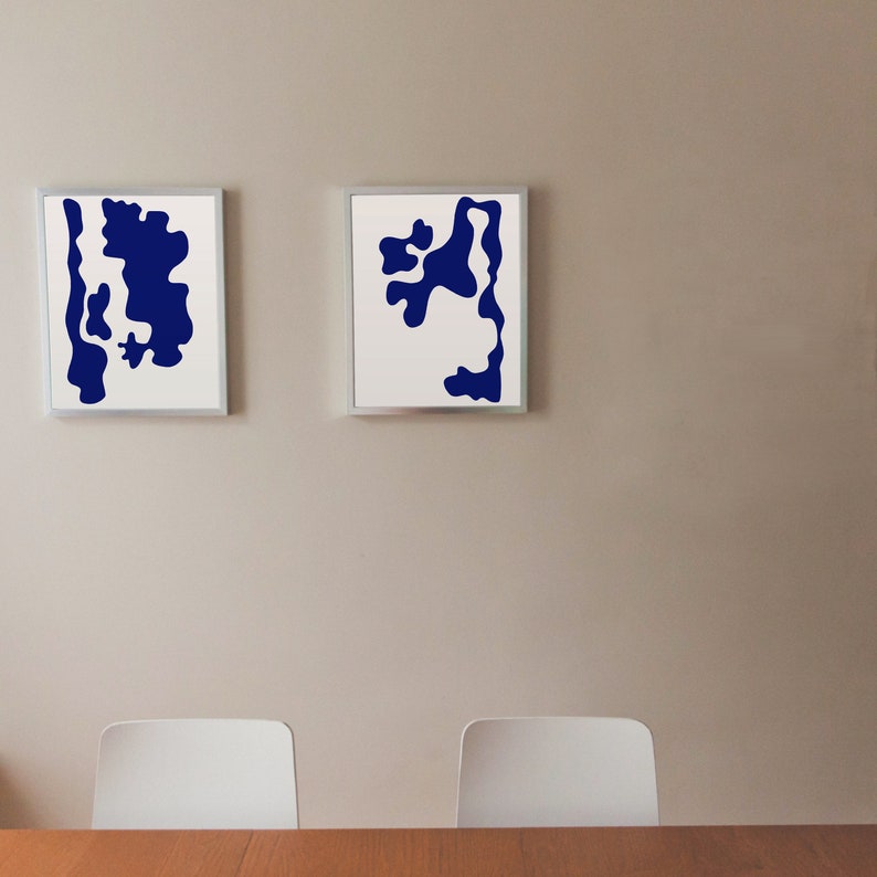 May include: Two framed abstract art prints with white backgrounds and blue abstract shapes. The prints are hanging on a light beige wall.
