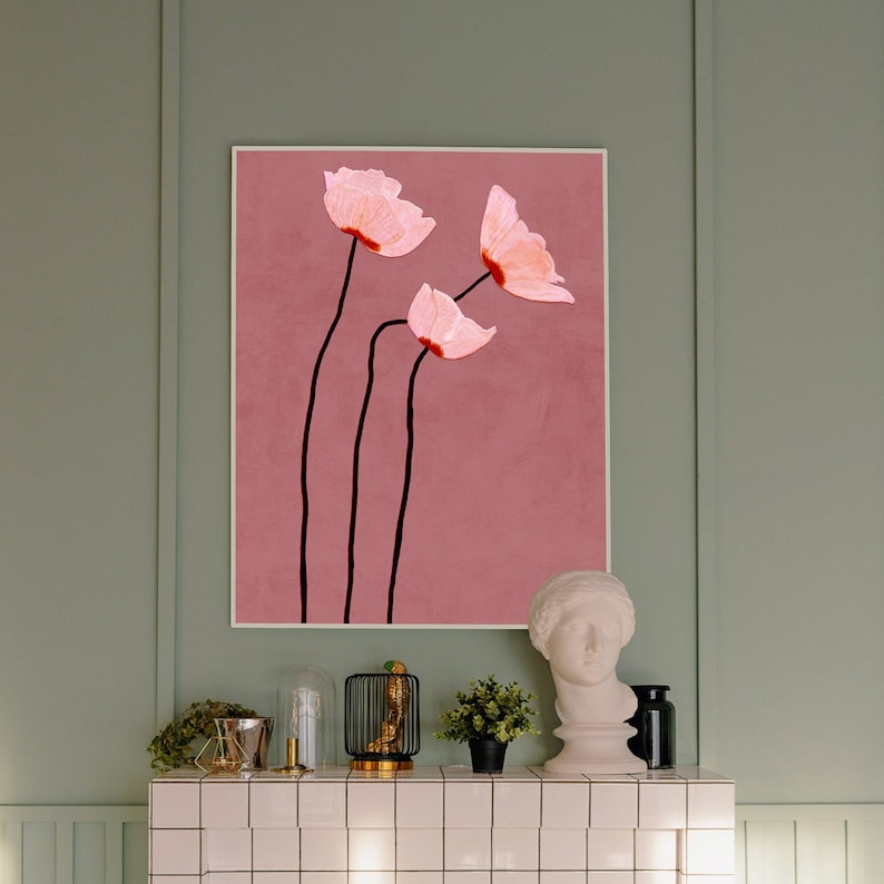 May include: A framed floral art print featuring three pink flowers with black stems against a dusty rose background. The artwork is displayed on a light green wall above a white tiled surface, with decorative items like a bust and small plants.