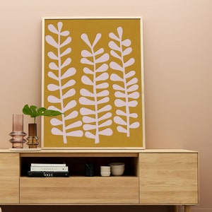 Op de afbeelding: Een ingelijst drukwerk met een mosterdgele achtergrond en roze gestileerde bladeren. De print hangt aan een lichtroze muur boven een houten kast met een plank en lades.