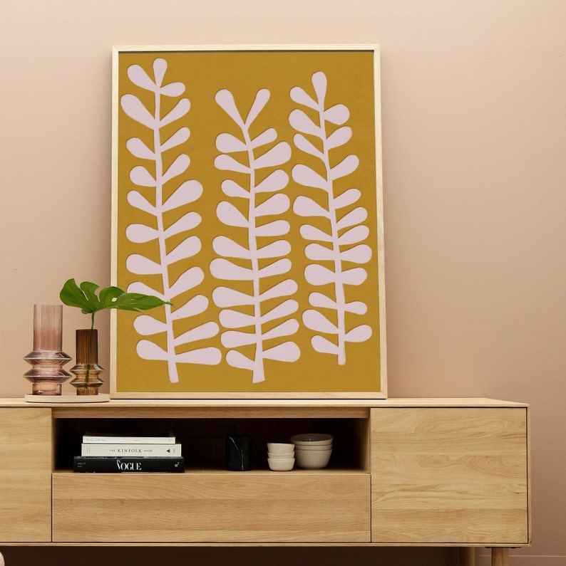 Op de afbeelding: Een ingelijst drukwerk met een mosterdgele achtergrond en roze gestileerde bladeren. De print hangt aan een lichtroze muur boven een houten kast met een plank en lades.