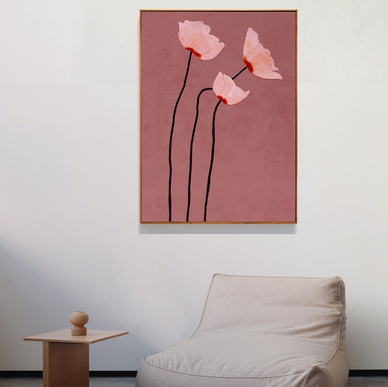 May include: A framed abstract floral art print. The artwork features three pink flowers with black stems against a dusty rose background. The flowers have a textured appearance, and the frame is a light brown color.