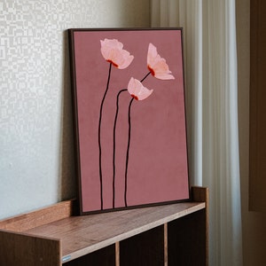 May include: A framed floral art print featuring three stylized pink flowers with red accents and black stems against a muted rose background. The artwork is displayed on a wooden shelf, adding a touch of nature-inspired decor.