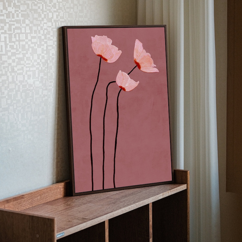 May include: A framed floral art print featuring three stylized pink flowers with red accents and black stems against a muted rose background. The artwork is displayed on a wooden shelf, adding a touch of nature-inspired decor.