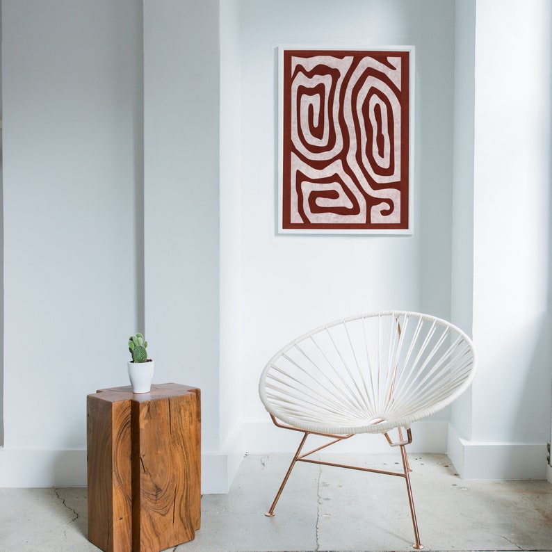May include: A white chair with a copper frame sits in a room with white walls. A wooden side table with a small potted plant sits next to the chair. A framed abstract print with a red and white design hangs on the wall.