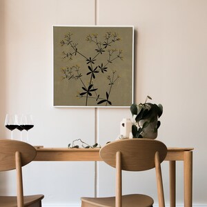 May include: A framed print of a black and white botanical illustration of a plant with yellow flowers on a beige background. The print is hanging on a wall above a wooden table with two chairs.