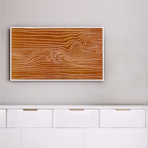 May include: A framed abstract print with white lines on a brown background. The print is hanging on a white wall above a white dresser with three drawers.