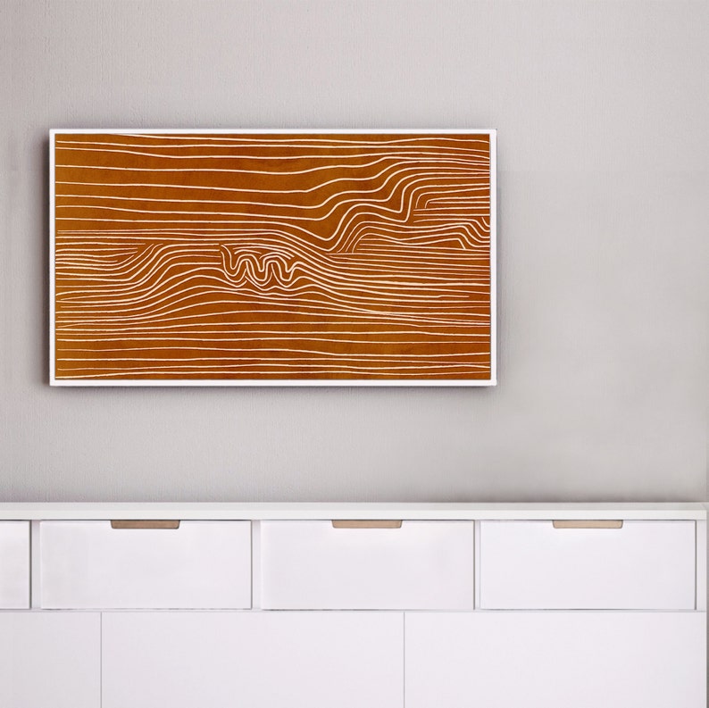 May include: A framed abstract print with white lines on a brown background. The print is hanging on a white wall above a white dresser with three drawers.