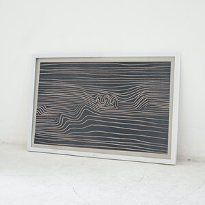 May include: A framed black and white abstract art print with a white frame. The print features a pattern of thin, wavy lines in a light brown color.