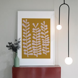 Op de afbeelding: Een ingelijst drukwerk met een mosterdgele achtergrond en roze gestileerde bladeren. De print hangt aan een witte muur boven een rood rek.
