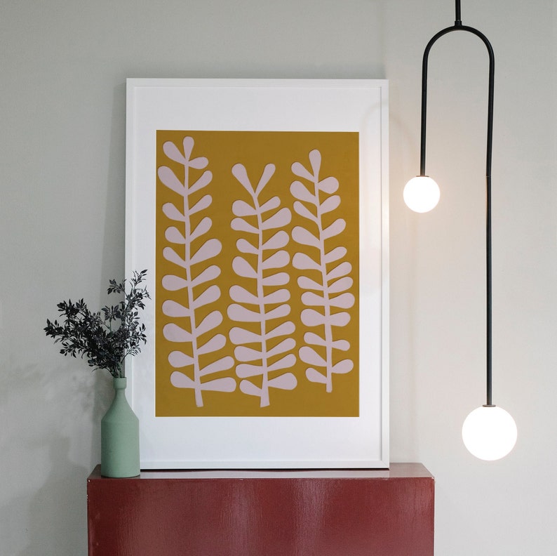 Op de afbeelding: Een ingelijst drukwerk met een mosterdgele achtergrond en roze gestileerde bladeren. De print hangt aan een witte muur boven een rood rek.