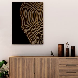 Puede incluir: Una impresi&oacute;n de arte de pared abstracta en negro y dorado con un patr&oacute;n de l&iacute;neas onduladas. La impresi&oacute;n est&aacute; enmarcada y colgada en una pared blanca sobre un gabinete de madera.