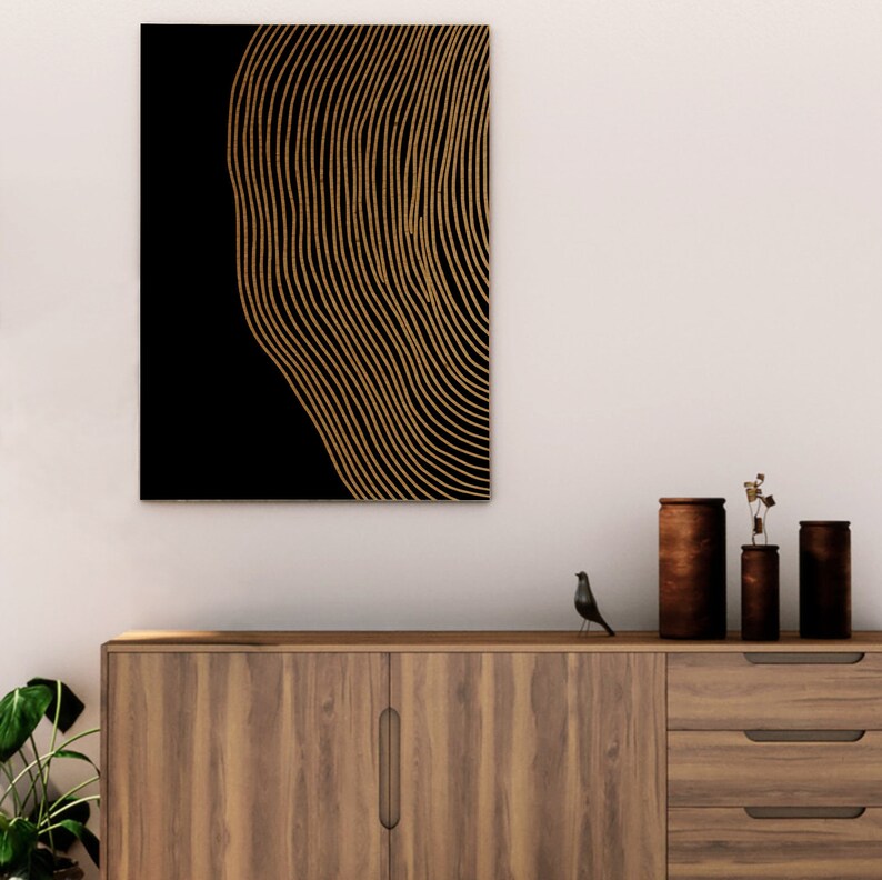 Puede incluir: Una impresi&oacute;n de arte de pared abstracta en negro y dorado con un patr&oacute;n de l&iacute;neas onduladas. La impresi&oacute;n est&aacute; enmarcada y colgada en una pared blanca sobre un gabinete de madera.