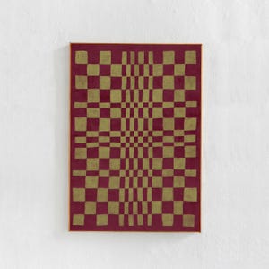 以下が含まれることがあります： 金とバーガンディの正方形が交互に配置された幾何学模様の額装アートワーク。 デザインは奥行きと動きの錯覚を生み出します。 アートワークは白い壁に展示されています。