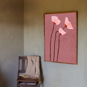 May include: A framed wall art print featuring three stylized pink flowers with black stems against a dusty rose background. The artwork is displayed in a room with a wooden chair and a beige throw.