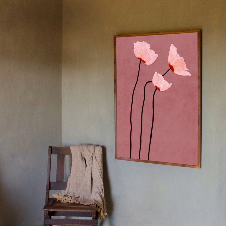 May include: A framed wall art print featuring three stylized pink flowers with black stems against a dusty rose background. The artwork is displayed in a room with a wooden chair and a beige throw.