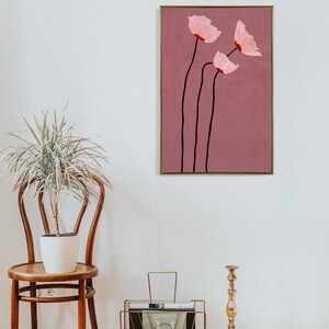 May include: A framed floral art print featuring three pink flowers with black stems against a mauve background. The artwork has a gold frame and is displayed on a white wall. The style is minimalist and modern, suitable for home decor.
