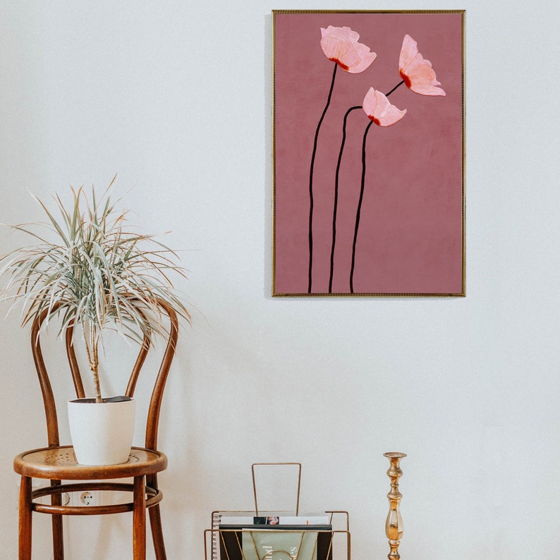 May include: A framed floral art print featuring three pink flowers with black stems against a mauve background. The artwork has a gold frame and is displayed on a white wall. The style is minimalist and modern, suitable for home decor.