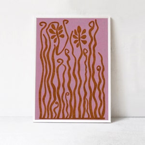 Op de afbeelding: Abstracte kunstprint met een roze achtergrond en bruine gestileerde bloemenvormen. De print is ingelijst in een wit frame.