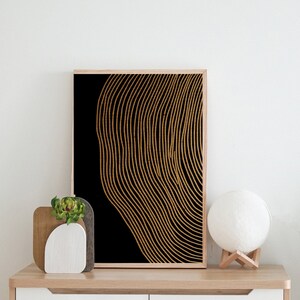 Puede incluir: Una impresi&oacute;n de arte abstracto en negro y dorado con un patr&oacute;n ondulado y rayado. La impresi&oacute;n est&aacute; enmarcada y colgada en una pared blanca sobre un estante de madera con una peque&ntilde;a planta y una l&aacute;mpara blanca.