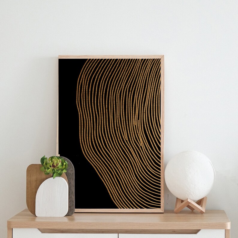Puede incluir: Una impresi&oacute;n de arte abstracto en negro y dorado con un patr&oacute;n ondulado y rayado. La impresi&oacute;n est&aacute; enmarcada y colgada en una pared blanca sobre un estante de madera con una peque&ntilde;a planta y una l&aacute;mpara blanca.