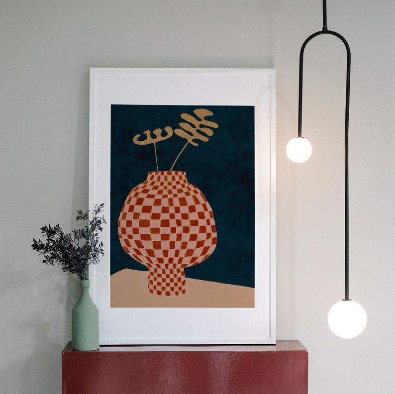 May include: A framed print of a stylized illustration of a red and white checkered vase with gold leaves on a dark blue background.