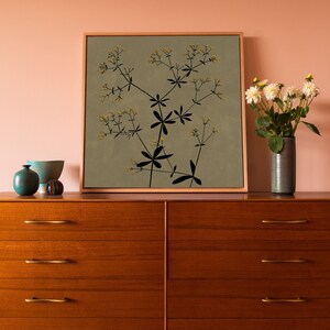 May include: A framed print of a botanical illustration featuring a black silhouette of a plant with small yellow flowers on a light green background. The print is displayed on a wooden dresser with two drawers.