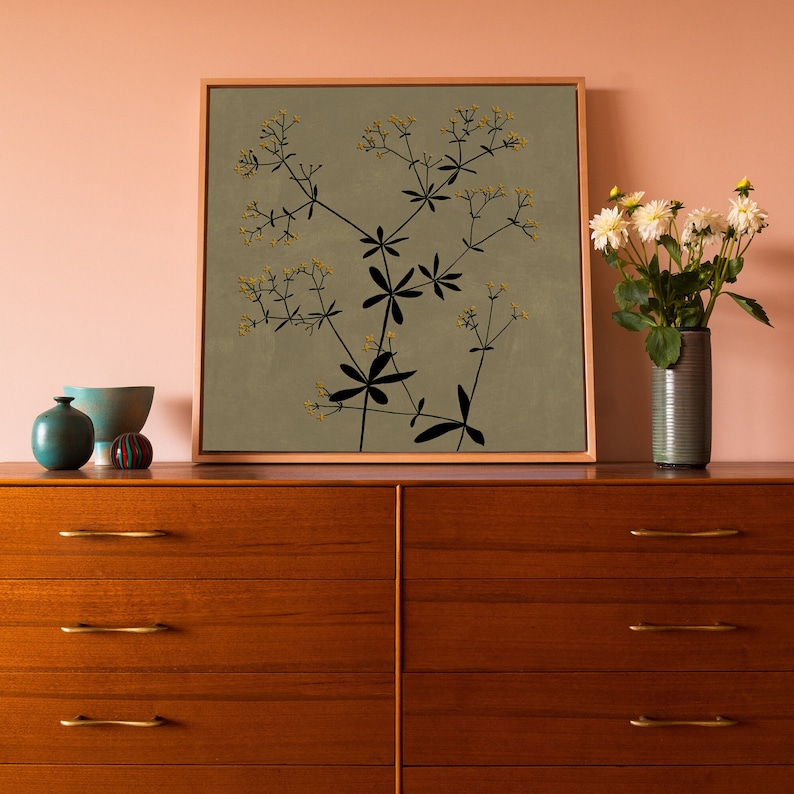 May include: A framed print of a botanical illustration featuring a black silhouette of a plant with small yellow flowers on a light green background. The print is displayed on a wooden dresser with two drawers.
