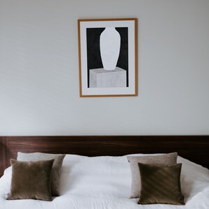 May include: A framed black and white print of a vase on a pedestal. The print is hanging on a white wall above a brown headboard with two brown throw pillows on a white bed.