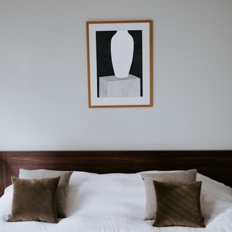May include: A framed black and white print of a vase on a pedestal. The print is hanging on a white wall above a brown headboard with two brown throw pillows on a white bed.
