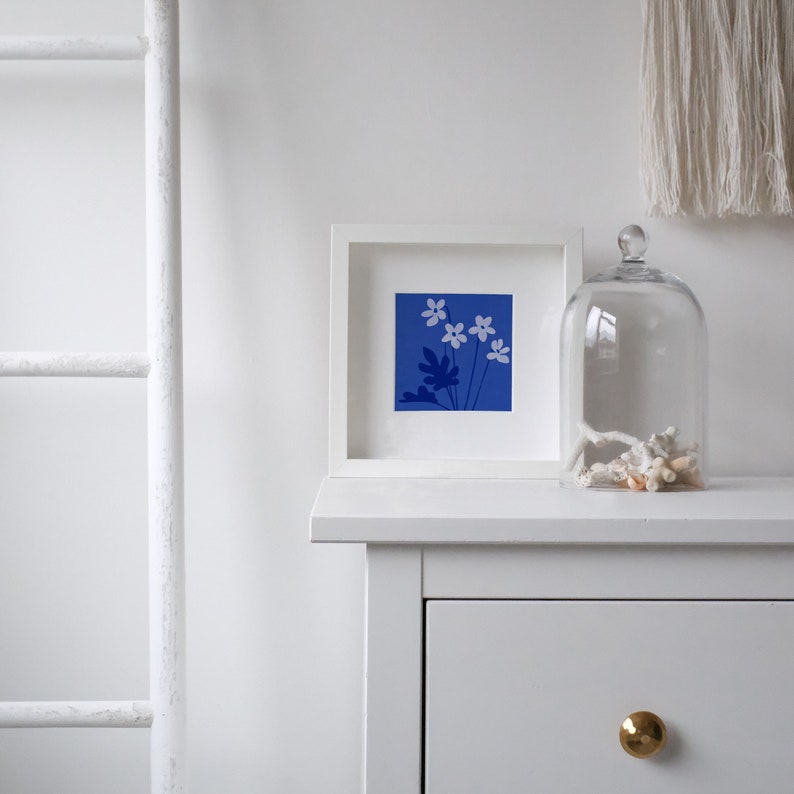 Pu&ograve; includere: Una stampa incorniciata bianca con uno sfondo blu e fiori bianchi. La stampa &egrave; appoggiata su una cassettiera bianca con un pomello dorato.