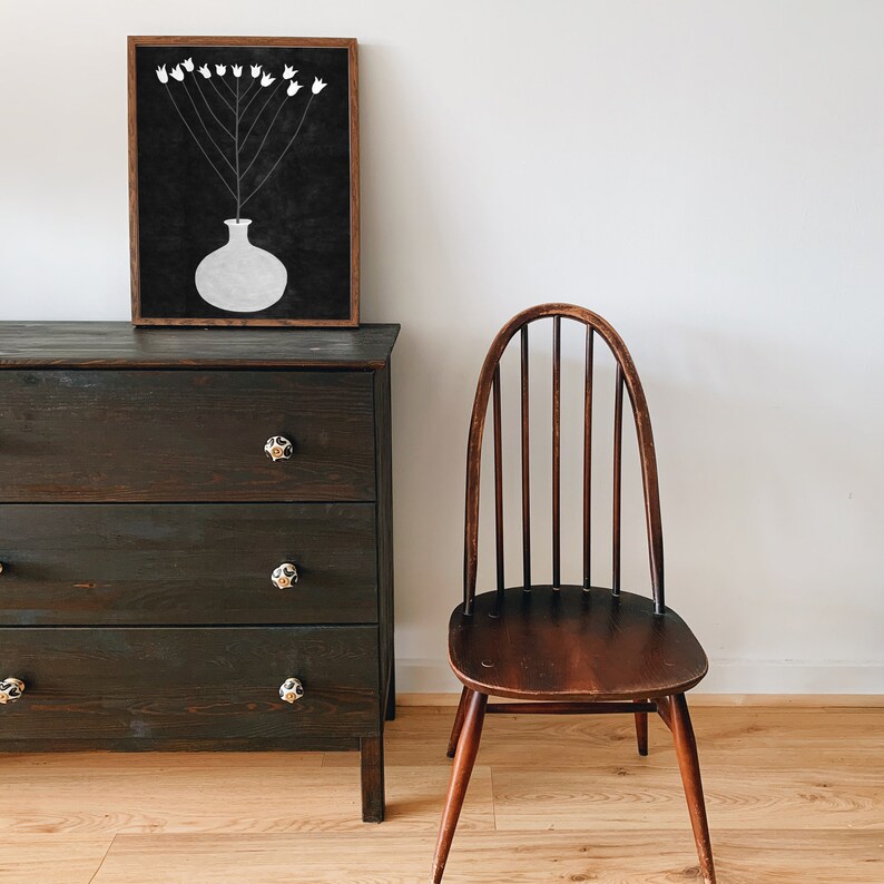 May include: A black framed print of a white vase with white flowers on a black background. A dark wood dresser with three drawers and a dark wood chair with a curved back are in the background.