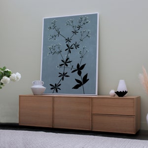 May include: A light wood cabinet with three drawers. The cabinet is in front of a light blue wall with a framed print of a black silhouette of a plant with white flowers.