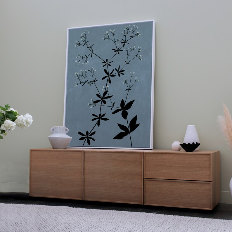 May include: A light wood cabinet with three drawers. The cabinet is in front of a light blue wall with a framed print of a black silhouette of a plant with white flowers.