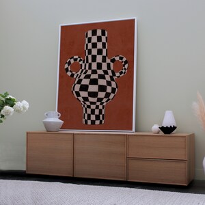 May include: A framed print of a black and white chequered vase with a red background. The vase is on a wooden cabinet with two drawers.