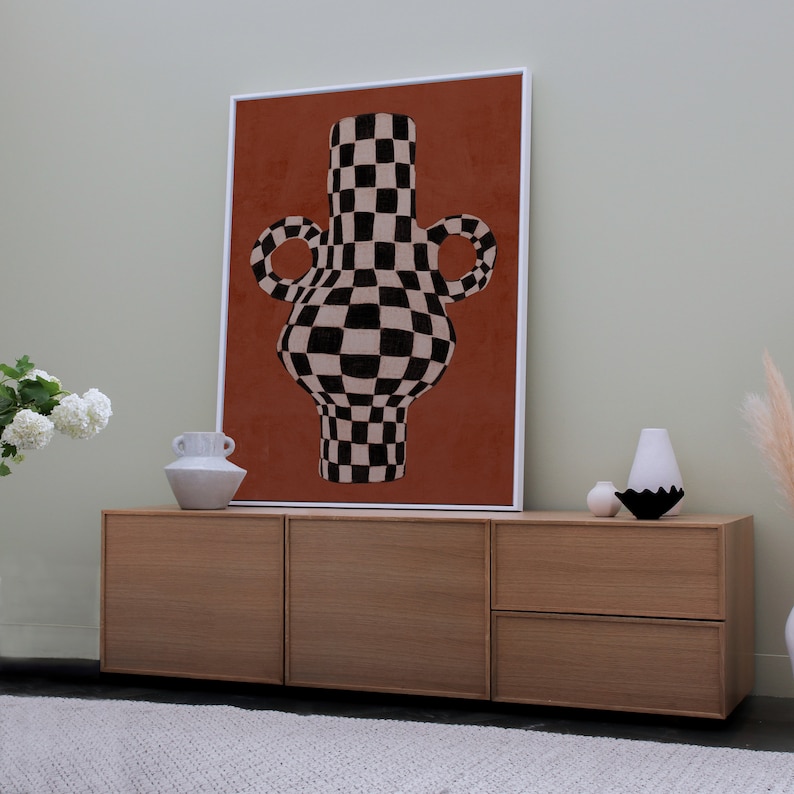 May include: A framed print of a black and white chequered vase with a red background. The vase is on a wooden cabinet with two drawers.