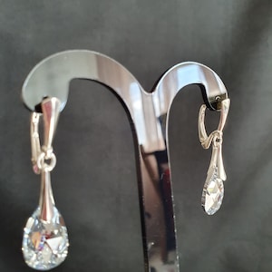 May include: A pair of silver teardrop earrings with clear crystal stones hanging from a black earring stand.