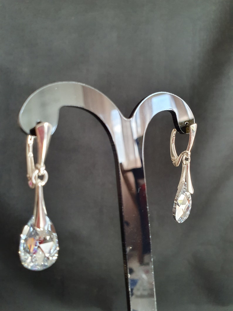 May include: A pair of silver teardrop earrings with clear crystal stones hanging from a black earring stand.