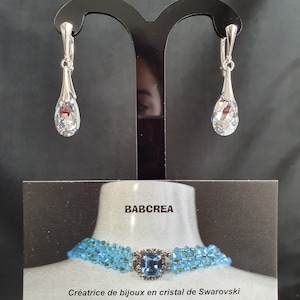 May include: A pair of silver teardrop earrings with clear crystal stones. The earrings are displayed on a black stand. The image also includes a business card with the text "BABCREA" and "Cr&eacute;atrice de bijoux en cristal de Swarovski".