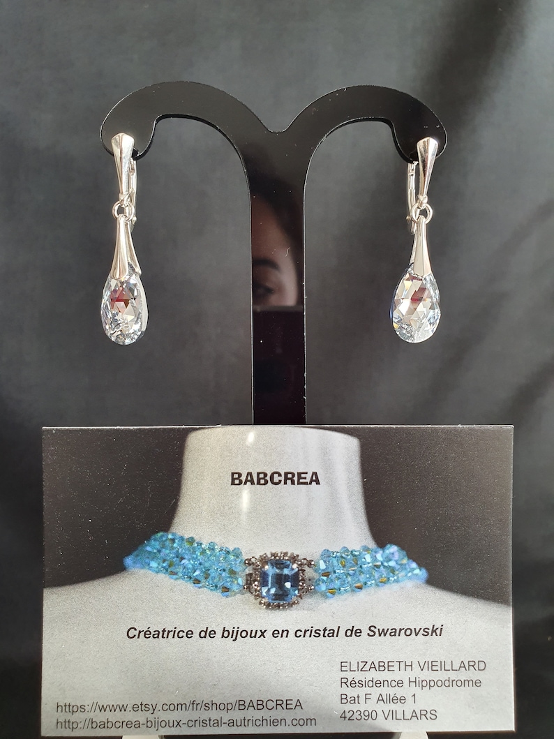 May include: A pair of silver teardrop earrings with clear crystal stones. The earrings are displayed on a black stand. The image also includes a business card with the text "BABCREA" and "Cr&eacute;atrice de bijoux en cristal de Swarovski".