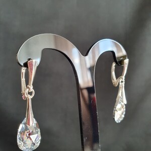 May include: A pair of silver dangle earrings with clear crystal teardrop-shaped stones. The earrings are hanging on a black display stand.