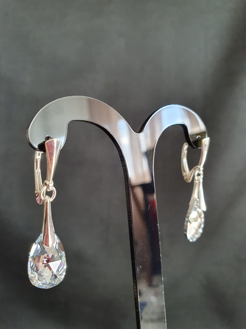 May include: A pair of silver dangle earrings with clear crystal teardrop-shaped stones. The earrings are hanging on a black display stand.