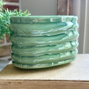 May include: A small, oval-shaped ceramic planter with a glossy, light green glaze. The exterior surface features a series of horizontal, undulating ridges that create a rippled texture around the entire circumference of the vessel.