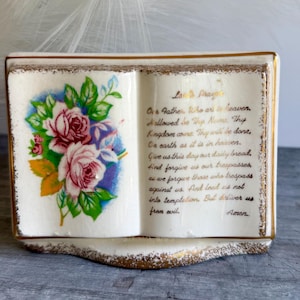Op de afbeelding: Een keramieken beeldje in de vorm van een open boek met een crèmekleurig glazuur en gouden rand. De linkerpagina bevat een kleurrijke illustratie van roze rozen met groene bladeren, terwijl de rechterpagina het Onze Vader in gouden cursief toont.