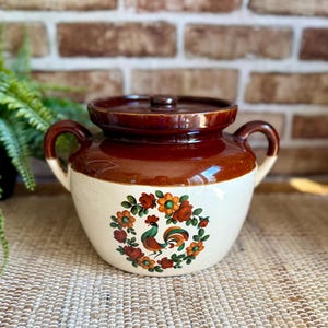 May include: A two-handled ceramic crock with a lid, featuring a cream-colored base and a dark brown glazed top. The front displays a colorful floral wreath design surrounding a central rooster illustration with red, orange, and green accents.