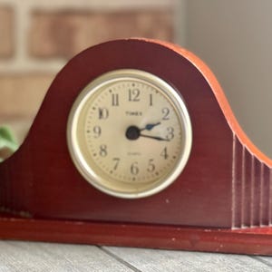 May include: A mantel clock with a dark reddish-brown wooden arched frame and a circular face. The clock features a cream-colored dial with black Arabic numerals, the Timex brand name, and thin black hands. The base is rectangular.