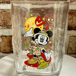 Può includere: Un bicchiere trasparente con una grafica colorata di Topolino vestito da safari con binocolo, circondato da coriandoli e una luna gialla. La base del bicchiere presenta in rilievo l'anno 2000 e il testo del copyright Disney.