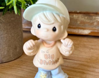 Może przedstawiać: Porcelanowa figurka dziecka w jasnej czapce z daszkiem, brzoskwiniowej bluzie z nadrukiem "I'm A Big Brother" w kolorze brązowym oraz jasnoniebieskich spodniach z małą żółtą łatką w okolicy prawego kolana.